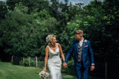 Charmaine and josh walking hand in hand in the grounds. The shot shows them from the knees up. they are holding hands and looking at each other. The bride is on the left and the groom is on the right. The bride wears a sleeveless, fitted, white dress. She has blond hair that she is wearing up. She has a bouquet in her right hand.