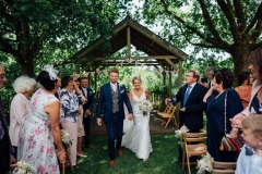 A full length shot of the couple walking away from the Oak Arbour after their ceremony. The couple are shown in full length with the arbour in the background. The groom is on the left and the bride is on the right. The groom wears a grey suit. The bride wears a full length, fitted, sleeveless , white dress and has a bouquet in her left hand