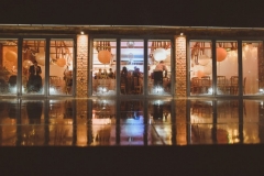 The Red Brick Barn. The picture is taken from outside at dusk. The full length glass windows are lit up and you can see guests relaxing inside