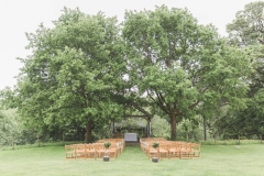 The Oak Arbour in May with each tree in full leaf. A full length shot showing the arbour set between 2 large oak tress. Wooden chairs are set out in the grass in frot of the arbour and the trees are in full leaf.