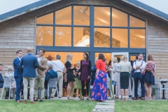 This picture shows the Green Room Bar from the outside with the building filling the full shot. The lights are on inside and it is dusk. The doors are open and guests are selling onto the lawn and patio in front. The building is an a fram with a central glazed panel. The rest is clad in oak.