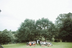 This is taken during an outdoor ceremony under the Oak Arbour. It is summer and the trees ae in ful leaf. The guests are seated in two colums of rows of chairs in front of the arbor with their abcks to the camera. The couple are under the arbour. There are some straw bales set out behind the last row of chairs. The image is taken from a distance with the Oak trees shown in full.
