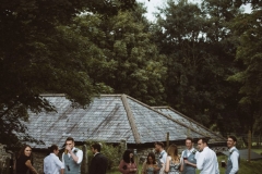 Some guests mingle in front of one of the cottages. You can seee the slate roof of the cottage in the background as the image is taken looking slightly down on the cottage. The guests are in the foreground and there are trees around the edges.