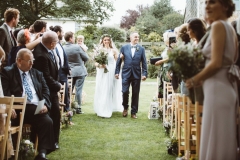 The bride walks down the \"aisle\" between the 2 colums of wooden chairs that are set out in front of the arbour. SHeis on her fathers arm. She is on the left walking towards the camera. She wears a full length white, sleeveless dress with lace details. She wears a veil. She has a bouquet of flowers, mostly green, in her right hand. The guests are stannding and turning around to see her.