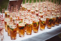 A shot of lots of glasses of Pimms waiting to get picked up by guests. The glasses are on a clothed white table. The glasses have Pimms and fruit in them and they have red, striped, paper straws.