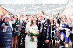 The bride and the groom stand side by side in the courtyard surrounded by their guests after their ceremony. The bride is on the left. She wears a fitted white gown with lace details and a veil. The pic is takes from the front the couple are standing surrounded by their guests who form a circle around them. The groom wears a dark suit with a white shirt and tie. The guests are throwing pom pom confetti at the bride and groom and the couple are laughing.