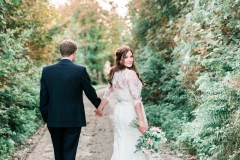 The bride and the groom are walking away from he camera down a narrow and leafy Cornish lane.  The bride is on the right. She wears a fitted white gown with lace details and a delicate floral crown. The pic is takes from the back and the bride is looking over her right shoulder back at the camera. The groom wears a dark suit. The bride holds a bouquet of white and pink flowers in her right and it is hand-tied.