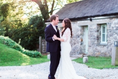 The bride and the groom are standing in front of cottage number 1. This cottage sleeps 2 people. It is made of local cob that is gray and brown in colour. it has a grey slate roof.  The bride and groom are standing in the middle of the shot and are facing each other and embracing. The bride is on the right. She wears a fitted white gown with lace details and a delicate floral crown. The groom wears a dark suit.