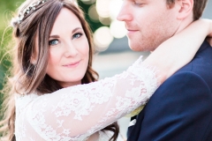 The bride and the groom are standing embracing.  This is a close up shot.  The bride and groom are facing each other and embracing. The bride is on the left and her head is turned towards the camera. She wears a fitted white gown with lace details and long sleeves and a delicate floral crown. The groom wears a dark suit and is looking at the groom.