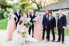 The bridal party are standing in front of cottage number 1. There group is made up of the bride and groom, 4 bridesmaids and 4 groomsmen. This cottage sleeps 2 people. It is made of local cob that is gray and brown in colour. it has a grey slate roof..  The bride and groom are standing in the middle of the shot and are standing side by side with the bride on the left.. She wears a fitted white gown with lace details and a delicate floral crown. The groom and groomsmen wear a dark suits. The bridesmaids are wearing full-length rose pink dresses with cap-sleeves.
