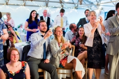 This picture is taken in the Red Brick Barn. It is of a group of wedding guests who are seated and standing in a group focussed on something that is out of shot. There are around 14 people in the group and they are taking pictures and smiling.