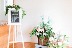 Decor details in the wedding barn, There is a white framed blackboard on an easel with an order of ceremony written on it. To the right of this there are some crates with florals in greenery, pink and white.