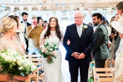 The bride and her father walk down the isle. The bride is on the left. She wears a fitted white gown with lace details and a veil. the pic is takes from the front of the wedding  barn and you can see bunting on the beams. The FOB wears a dark suit with a white shirt and a bow-tie. The bride holds a bouquet of white and pink flowers and it is hand-tied. There is a bridesmaid in the foreground to the left who holds a similar bouquet. The guests are standing bedside their chairs looking at the bride and FOG. Wooden folded chairs are set out in two columns of rows with an aisle down the middle.