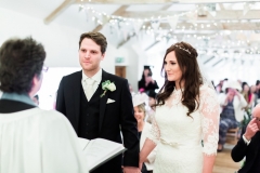 The bride and the groom stand in the wedding barn exchanging vows. The bride is on the right. She wears a fitted white gown with lace details and a veil. The pic is takes from the front of the wedding barn and you can see bunting on the beams. The groom wears a dark suit with a white shirt atie. T There is a priest in the foreground to the left who. The guests are seated looking at the bride and groom. Wooden folded chairs are set out in two columns of rows with an aisle down the middle.