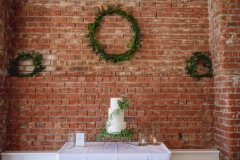 This is a picture of the cake table set up against the red brick wall in the Red Brick Barn. The shot shows quite a  lot of the wall and the cake is quite small in comaprison near the bottom of the image. The cake is simple, whie and 3 tiered , decorated with green leaves.  It is placed on a white-clothed table but only the table top. On the wall behind the cake