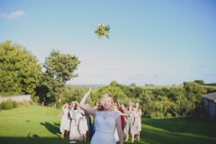 The bouquet toss. The bride is srtanding facing the camera with a group of female guests gathered behind her. The sky is bright blue and the shadows are long indicating that is it early evening. Everyone is standing on a green lawn and there are trees in the backgound in full leaf. The bride has her eyes squeezed shit and is throwing her bouquet over her right shoulder, as hard as she can, with her right arm. The bride wears  a sleeveless, white dress with lace details  and a beaded bodice.