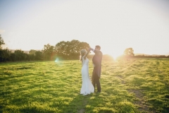 This is an image of the bride and groom standing in a field at sunset. . They are standing in the middle of the picture. The sun is setting behind them and there is a row of green treesin the background.  The groom is wearing a navy suit, gray waistcoat, white shirt and blue tie. The bride wears  a full-length, sleeveless, white dress with lace details  and a beaded bodice.
