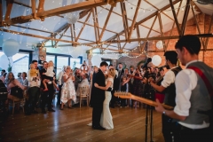 This is an image of the Red Brick barn during a wedding reception. The room has oak floors and a red brick wall and white ceiling. There are people grouped in the far right hand corner of the room in front of a set of bi-fold doors watching the bride and groom having their first dance. In the foreground is a musician playing a guitar with his back to the camera.  There are balloons rising up towards the A-frame beams in the pitched roof.  The room has oak floors and white walls and ceiling. The tables are round and covered in white table cloths. There are bi-fold doors to the front of the room away from the camera. Light is streaming in the windows. The lights are dimmed and the couple are dancing closely in the middle of the floor.