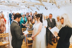 Amy and Kieran exchanging vows at the front of the wedding barn with their guests seate behind them.