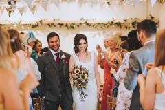 Amy and Kieran  leaving the weding barn hand in hand and smiling being cheered by their guests. Amy holds a bouquet of sunflowers