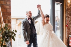 The bride and groom are walking into the Red Brick barn. They are holding hands and have their hands raised in celebraition. The bride has She has dark hair worn up. She has a fitted white dress with a sweatheart neckline and lace sleeves. The groom is wearing a tweed 3 piece suit and brown bow-tie.