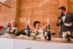 The wedding party are at their places at a long "top" table in front of the Red Brick wall in the barn. The best man is on hos feet making a speech and the bride has her arms lifted in a cheer,