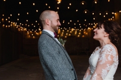 The couple are standing inside the Really Rustic Barn.  You can see festoon lighting in the backgorund inside the barn. The are facing each other and the shot is fromthe waist up.  The bride has She has dark hair worn up. She has a fitted white dress with a sweatheart neckline and lace sleeves. The groom is wearing a tweed 3 piece suit and brown bow-tie.