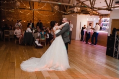 The couple are having their first dance in the Red Brick Barn. You can see some guests and a bar in the backgorund inside the barn. The are facing each other side on to the camera and embracing.  The bride has She has dark hair worn up. She has a fitted white dress with a sweatheart neckline and lace sleeves. The groom is wearing a tweed 3 piece suit and brown bow-tie.