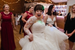 The bride is dancing and swirling her skirt  in the Red Brick Barn. You can see some bridesmaids and a bar in the backgorund inside the barn. She is facing the camera and smiling broadly.  The bride has She has dark hair worn up. She has a fitted white dress with a sweatheart neckline and lace sleeves and a full skirt..