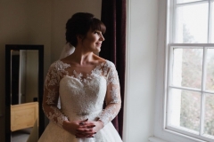 A bride faces the camera, her head is turned to lookout of a window to her left. She has dark hair worn up. She has a fitted white dress with a sweatheart neckline and lace sleeves