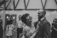 The couple are dancing in the Red Brick Barn. You can see some guests and in the backgorund inside the barn. The are facing each other side on to the camera and embracing.  The bride has She has dark hair worn up. She has a fitted white dress with a sweatheart neckline and lace sleeves. The groom is wearing a tweed 3 piece suit and brown bow-tie.