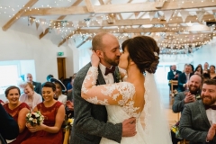 The bride and groom are facing each other and embacing in the wedding barn during their ceremony. The bride has She has dark hair worn up. She has a fitted white dress with a sweatheart neckline and lace sleeves. The groom is wearing a tweed 3 piece suit and brown bow-tie.