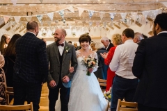 The bride and groom are walking out of the weddin barn holding hands after their ceremony. The bride has She has dark hair worn up. She has a fitted white dress with a sweatheart neckline and lace sleeves. The groom is wearing a tweed 3 piece suit and brown bow-tie.