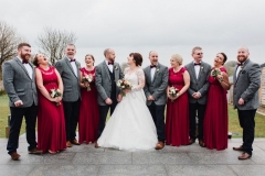 An informal group shot of the bride and groom and their 4 bridesmaids and groomsmen. The bridesmaids they are wearing red dresses and holding flowers. The bride has dark hair worn up. She has a fitted white dress with a sweatheart neckline, lace sleeves and a full skirt. The groom is wearing a tweed 3 piece suit and brown bow-tie. the groomsmen are dressed like the groom.