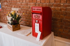 There is a white clothed table in this picture with a red brck wall in the background. On the table is a red post box (used for cards). There is also a box of flowers.