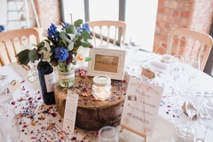 This picture shows a table centrepiece made up of a log slice, a candle in a lace jar, a table name and table confetti.