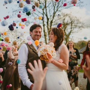 This image shows a bride and groom outside with some other guests standing around them. The guests are throuwing pompom confetti. The couple are in the foreground and the shot is from the knees up. The couple are standing side-by-side with the groom on the left and the bride for the right.The bride has dark hair which she is wearing loose. She's wearing an a-line sleeveless dress with the lace detailing at the shoulders. In her left hand she carries a bouquet of muted pink and white peonies. The groom has dark hair and is wearing a white shirt and a brown tweed waistcoat. The groom has his eyes closed and is smiling the bride is looking at her groom and laughing. In the background there are some trees without leaves. In the foreground there is a hand throwing confetti.
