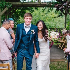 Newly weds leaving the Oak Arbour. The arbour is a wooden structure with four corner posts supporting a roof. This photo shows a couple just walking away from the Oak Arbour after their ceremony. The couple are in the foreground and the shot shows them from the knees up. They are holding hands and the grrom is on the left, the bride on the right. You can see some guests around then. The arbour is decorated with flowers and greenery. There are some chairs visible in the background in the arbour. The trees in the background I in full leaf. The bride has dar hair which she wears loose. She is wearing a white, fitted sweetheart neckline dress with lace overlay and has a veil. In her left hand she is holding a bouquet of white and purple flowers. The groom is wearing a three piece mid blue suit with white shirt and a patterned bow-tie. The couple are smiling and looking at the camera.
