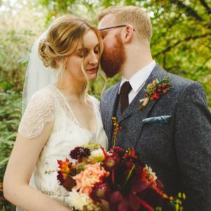 This is a close up photo of a bride and groom embracing in the woods. The bride is wearing a cream dress with lace detail and lace sleeves she is wearing a veil. She has dark blonde here which she is wearing in a loose updo. The groom is wearing a grey, tweed three-piece suit with a white shirt and a brown tie. The groom has glasses and a full beard and he has red hair. In her right hand the bride is holding a bouquet with autumn coloured flowers; purple red and yellow. The bride is on the left of the shot and the groom is on the right. The bride is turned slightly towards the groom and has her eyes closed. The groom is on the right and is leaning in towards the bride and is kissing her on the far side of her head. The bride has her eyes closed.