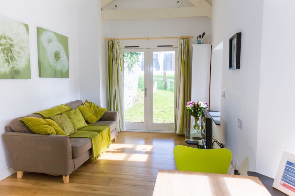 This is a photo of the lounge in cottage number one which is our smallest cottage that sleeps two people. The photograph is taken looking towards the French windows in the room on the right hand side of the cottage. The window looks out on to the grounds. At the top of the photograph you can see the A-frame beams in the roof of the cottage these are painted white. All the walls are also painted white. On the French windows there are some green white striped curtains. On the left-hand wall there are two pictures showing dandelion clocks. There is an oak floor. On the left hand side there is brown sofa with lime green cushions (five) and a lime green throw. On the right hand wall there is a television and vase of pink and white flowers. On the right-hand wall there is a black framed picture. In the foreground is a small dining table with a line green chair. There is one window visible on the right and there is slate windowsill in this window. In the far right hand corner you can see the side of a tall bookcase.