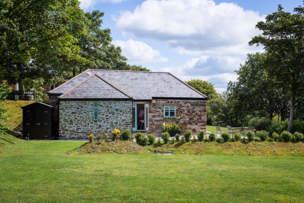 This picture shows cottage number two which is a detached cottagethat sleeps 4 people. The cottage is in the middle of the image and there are lawns in front and a blue cloudy sky above. There are trees in leaf to the left and to the right. To the left of the cottage there is a small black shed. In front of the cottage is a slightly raised bank with a small box hedges planted along top. The cottage is built of local granite stone which is brown and grey in colour. It has a grey slate roof the main roof line runs from left to right along the length of the photograph. In the middle of the building there are some French doors one is open. The doors are painted pale blue on the outside. There is a long narrow window painted blue on the left of the door. On the right of the door there is a georgian style sash window also painted blue.