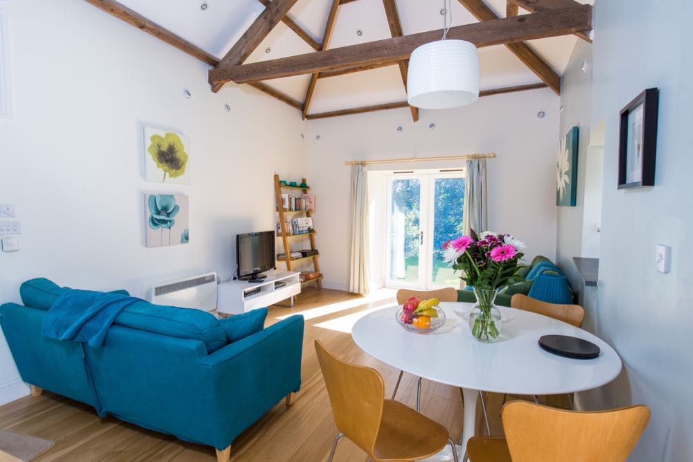 This image is of the lounge/dining area of cottage number two which is a sleeps four detached cottage. The room has oak flooring. There is a vaulted ceiling with many old oak a-frame beams supporting the roof. The walls are painted pale blue and the ceiling is white. There are spotlights in the roof and around the edge of the ceiling. There is a two seater blue sofa, a white TV table with a TV on top of it, a ladder shelving unit with five shelves. There is a second two seater sofa that is green. The dining area to the right of the photo has a round white pedestal table and four wooden dining chairs. On the dining table there is a vase of flowers and a bowl of fruit. On the wall to the left there are two prints one above the other line green and a blue floral design. There are two pictures on the right-hand wall one is blue with a floral design. There is a pendant light above the table.