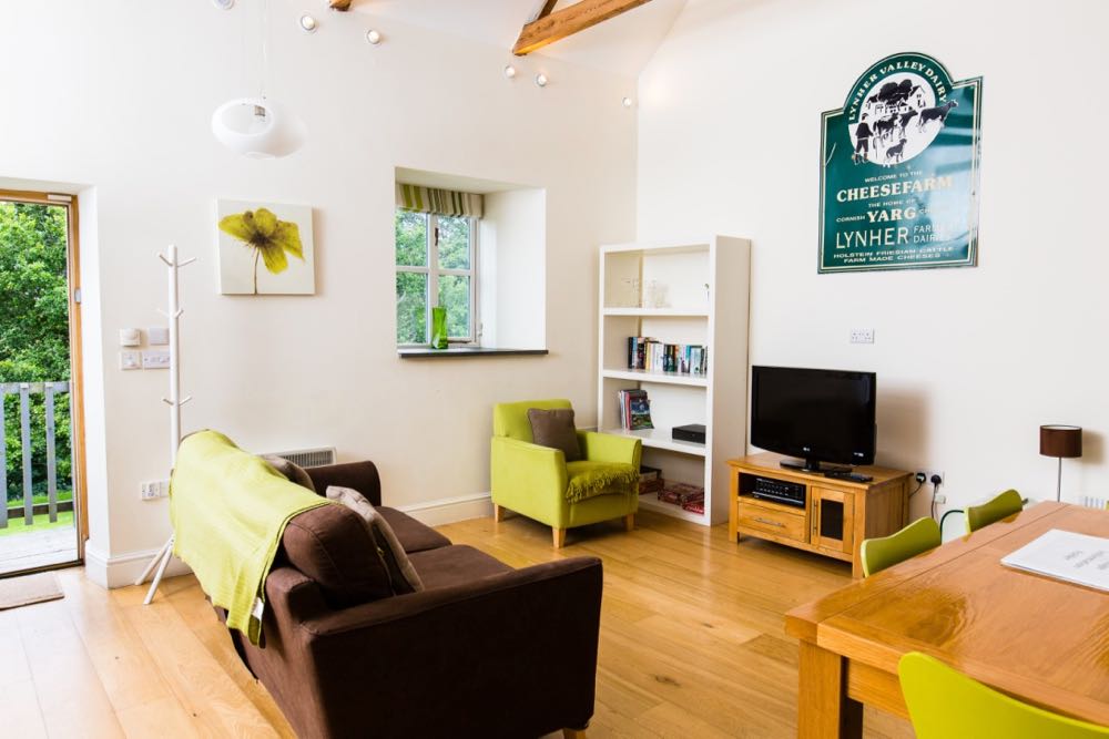 This is an image of the lounge area of cottage number three which is a sleeps 4 cottage. The room has oak flooring. The front door can be seen and the door is open. OUtside the door you can see some wooden ballustrades and green trees in leaf. The walls are all painted cream. At the top of the wall you can just see the edge of the a-frame beams that support the roof. There are spotlights around the edge of the ceiling. There is a pendant light hanging to the right of the door. There is one window which is a casement style four pane window with a striped blind that is open. Out of the window you can see more trees. There is a slate windowsill. There is a wooden tree-style coat rack. There is a brown sofa with a lime green throw. There is a lime green armchair with a brown cushion. There is a white bookcase with four shelves. There is an oak TV unit with a TV on top. On the wall above the television there is an old advertising sign for a cheese farm which is green with white writing. To the right of the photo you can see an oak dining table and three of the four lime green dining chairs.