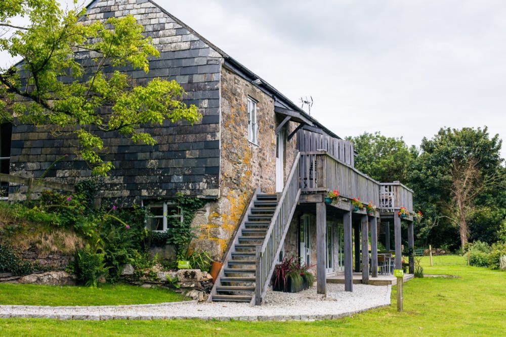 This is an exterior shot of a large barn that is split into three cottages namely cottage three, cottage four and cottage five. The barn has two storeys. Cottage three and cottage four are upstairs. Cottage five is on the ground floor. In the image the large barn is seen from the side. The side of the barn is covered in grey hanging slate. There is a window on the bottom right hand corner. This is a window of cottage 5. The window is a four paned casement style window and is painted pale gray. To the front of the building (which is on the side of the photograph) there is a set of grey painted wooden steps leading upwards to the second floor of the building. There is a balcony along the full length of the second floor. The balcony is painted grey and is made of wood. The balcony is supported on wooden posts from the ground. The area immediately around the cottage is a gravel path and at the far end of the front of the building you can see a decking area with some outside chairs and a table. All around the gravel path is lawn and to the left and to the right are trees in the leaf. The sky is overcast. To the front of the building on the first floor you can see a casement style four pane window nd a door both are painted gray. This is the window and the door of cottage number three. On the ground floor of the front of the building you can see two sets of french windows and the front door all are painted gray. These are the french doors and the front door of cottage number five.