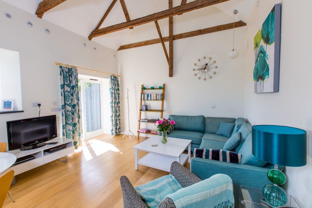 This is an image of the lounge area in cottage number four which is a sleeps four cottage. The cottage has oak flooring. All the walls are painted cream. The ceiling is painted white. You can see three of the large, old, oak, a-frame beams which support the vaulted roof. In the left-hand corner of the photo you can see the edge of the dining table which is white and the edge of two of the wooden dining room chairs. You can see the edge of the window reveal and the slate window sill. There are spotlights around the edge of ceiling. There are French doors and around the French there is a set of white curtains with a blue floral design. There is a white gloss TV unit with a television on top. The curtain rail is made of walk. There is a tree style coat rack. There is a ladder style bookcase with five shelves. The bookcase is made of oak. There is a small, round, white pendant light in the corner of the room. There is a large feature clock which is a spoke and design in cream with 12 balls and black hands. There is a large picture with teal and lime flowers. There is a large teal corner sofa with two teal cushions and a navy and teal striped cushion. There is a patterned grey and teal armchair with a teal and white striped throw. In the right-hand corner of the image you can see a glass side table with a table lamp with a green and teal glass ball design and a teal shade.