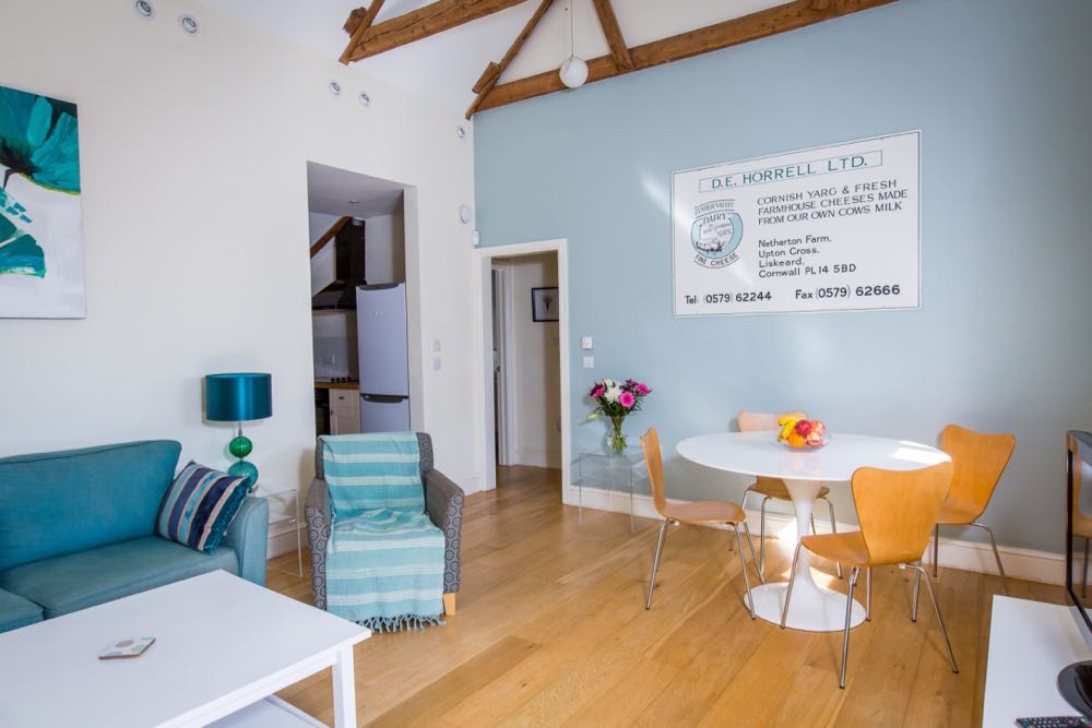 This is an image of the dining/lounge area in cottage number four which is a sleeps four cottage. The cottage has oak flooring. All the walls are painted cream. The ceiling is painted white. The wall in front of the camera is painted pale blue. There is a door on the left leading to a kitchen. There is a door to the right of that door leading into a corridor. There is an old advertising sign for the cheese farm on the blue painted wall. The sign is white with teal and black font. You can see the edge of 2 of the large, old, oak, a-frame beams which support the vaulted roof. In the middle of the photo you can see the the dining table which is white and round with a pedestal. There are 4 wooden dining room chairs. There are spotlights around the edge of ceiling. In the bottom right hand corner of the image you can see the corner of a white gloss TV unit made of walk. TTo the left of the table there is a glass table with a vase of pink and white flowers on it. There is a small, round, white pendant light in the corner of the room. There is a large picture with teal flowers on the wall on the left. There is a large teal sofa with a navy and teal striped cushion. There is a patterned grey and teal armchair with a teal and white striped throw. To the right of the sofa you can see a glass side table with a table lamp with a green and teal glass ball design and a teal shade. There is a white painted wooden coffee table in front of the sofa.