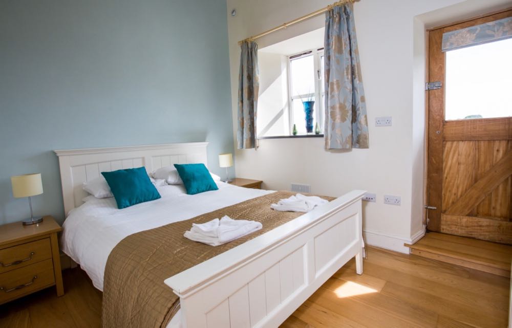 This image is of the double bedroom in cottage number four. The room has oak flooring the wall behind the bed is painted pale blue. There are two oak bedside tables one on each side of the bed. Each has two drawers. On each bedside table there is a stainless steel bedside light with a cream shade. The bed is made of wood and is painted white. The bed linen is white there are four pillows two on each side. On each set of pillows there is a teal cushion. At the base of the bed there is a folded gold throw. On this throw there are two sets of folded white towels. The wall right of the bed is painted cream in on this wall there is a door and a window. The window is a four pane casement style window there is a slate windowsill. On the slate windows ill there is a blue vase. The curtains are pale blue with a gold floral design. The door leads to the outside. The door is a half glazed door there is a blind with the same fabric as the curtains on the glazed section of the door. The door is made of oak. There is a step from the floor level to exit the door.