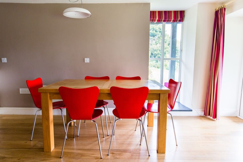 This image is of the dining area of cottage number five. Cottage number five sleeps six people. The image shows a rectangular dining table which is made of oak. Around the table there are six red dining chairs. The floor is made of oak. The wall behind the table is painted at taupe colour. There is a white bowl shaped pendant light above the table. To the right of the table there is a large full height four pane casement window. This window goes from ceiling to floor. There is a slate windowsill. There is a striped blind in red cream and brown tones. In the wall to the right of the photo there is another window you can see a full length curtains in this window made of the same fabric as the blind.