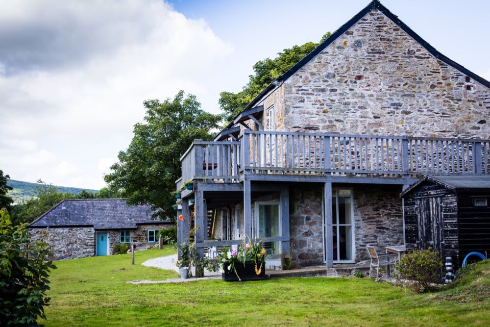 This is an exterior shot of a large barn that is split into three cottages namely cottage three, cottage four and cottage five. The barn has two storeys. Cottage three and cottage four are upstairs. Cottage five is on the ground floor. In the image the large barn is seen from the side. The side of the barn is made of local granite cob in gray and brown. It has a pitched roof with a gable end. There is a window on the bottom left hand corner. This is a window of cottage 5. The window is a four paned casement style window and is painted pale gray. To the front of the building (which is on the left side of the photograph) there is a set of grey painted french windows (these lead into cottage 5). There is a balcony along the full length of the second floor. The balcony is painted grey and is made of wood. The balcony is supported on wooden posts from the ground. The area immediately around the cottage is mostly lawn with a gravel path in the perimeter immediately around the cottage. In the left hand corner of the picture there is a hill sloping gently off to the left. Cottage number 2 can be sen in the backgound. This is a detached cob cottgae with gray slate roof and a blue painted door and windows. In the backgound there are some trees leaf. The sky is overcast.