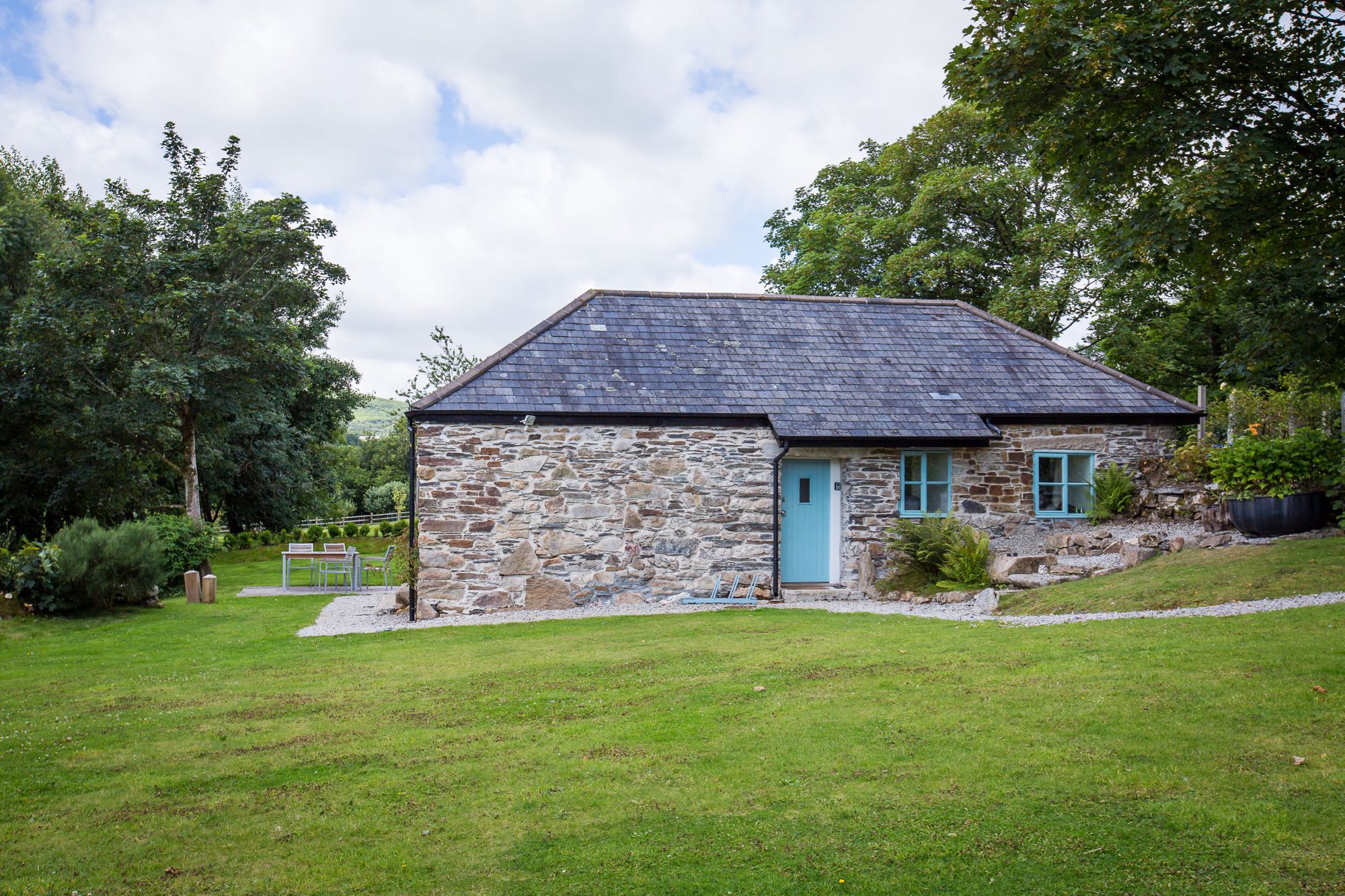 This picture shows cottage number two which is a detached cottage that sleeps 4 people. The cottage is in the middle of the image and there are lawns in front and a blue cloudy sky above. There are trees in leaf to the left and to the right. The cottage is built of local granite stone which is brown and grey in colour. It has a grey slate roof the main roof line runs from left to right along the length of the photograph. In the middle of the building there is the front doorthat is painted pale blue. On the right of the door there are 2 georgian style sash window also painted blue.